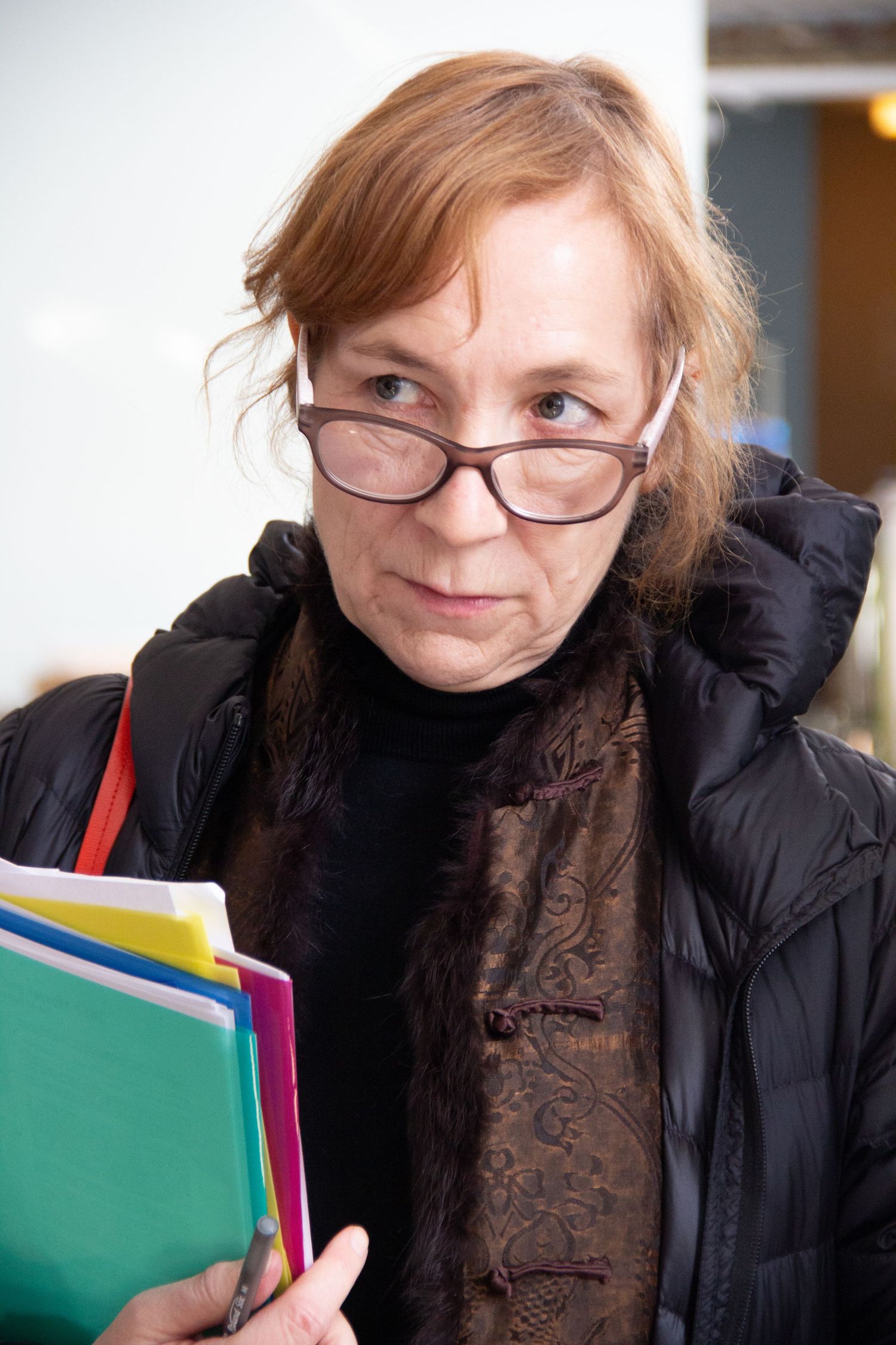 Middle-aged woman with red hair and glasses holding colorful folders, wearing a black puffer jacket indoors.