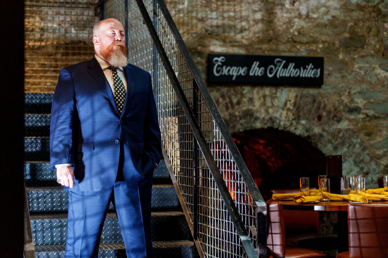 A bearded man in a blue suit stands by industrial stairs near an Escape the Authorities sign.