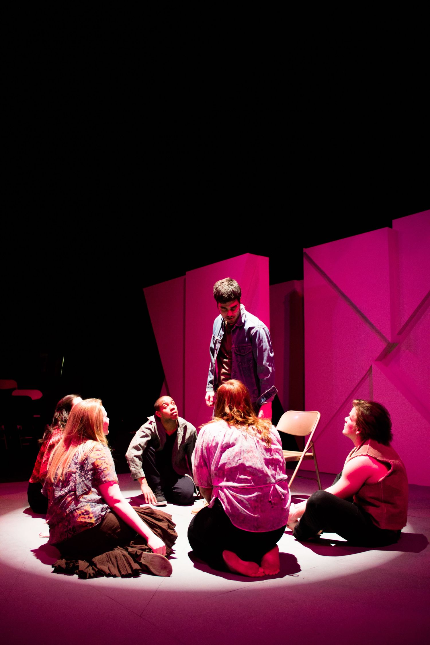 Performers seated on floor under dramatic pink spotlight during theatrical scene.