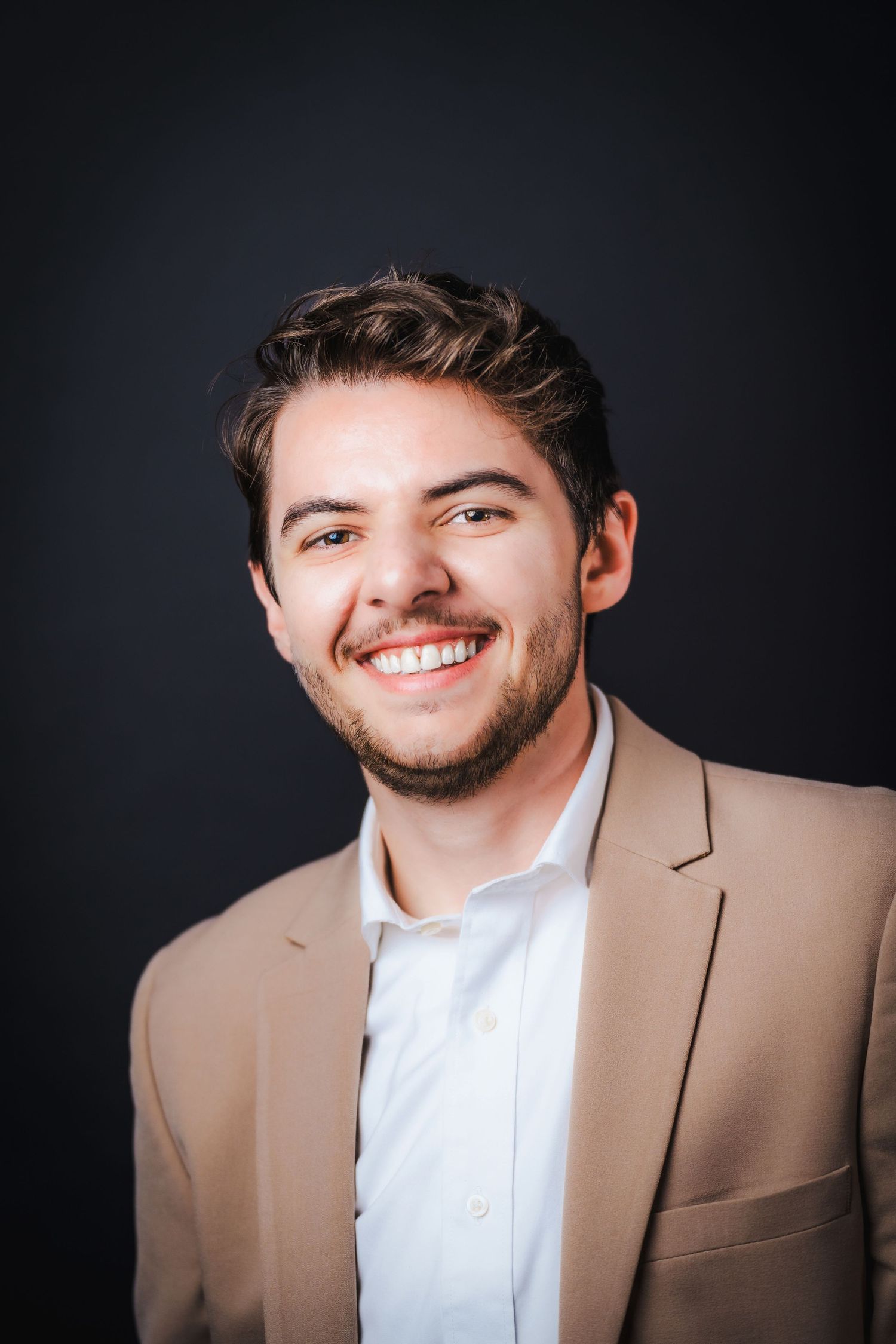 Professional headshot portrait set against dark background showing person in tan suit and white dress shirt.