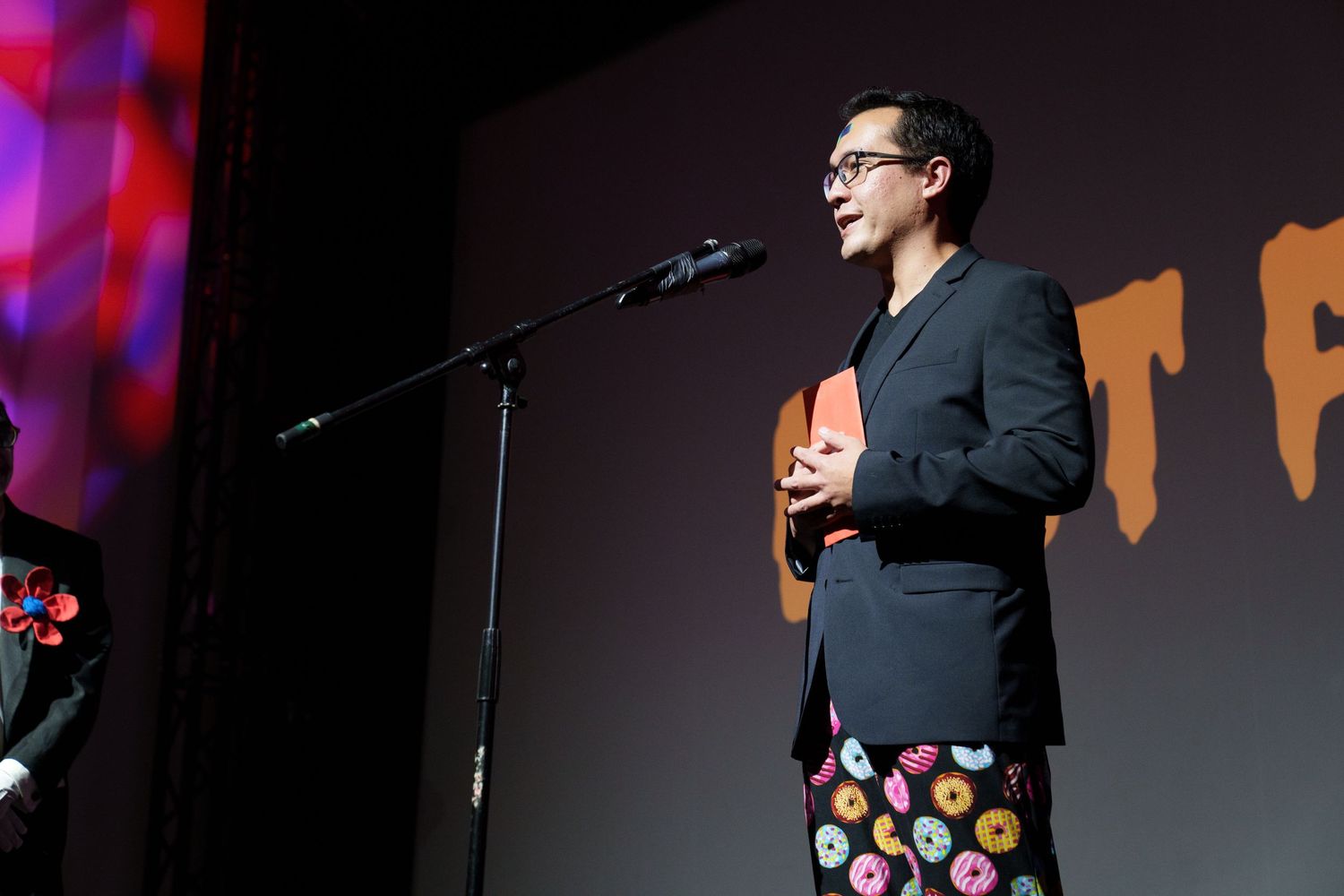 Stage performer in black attire against colorful backdrop with dramatic lighting.
