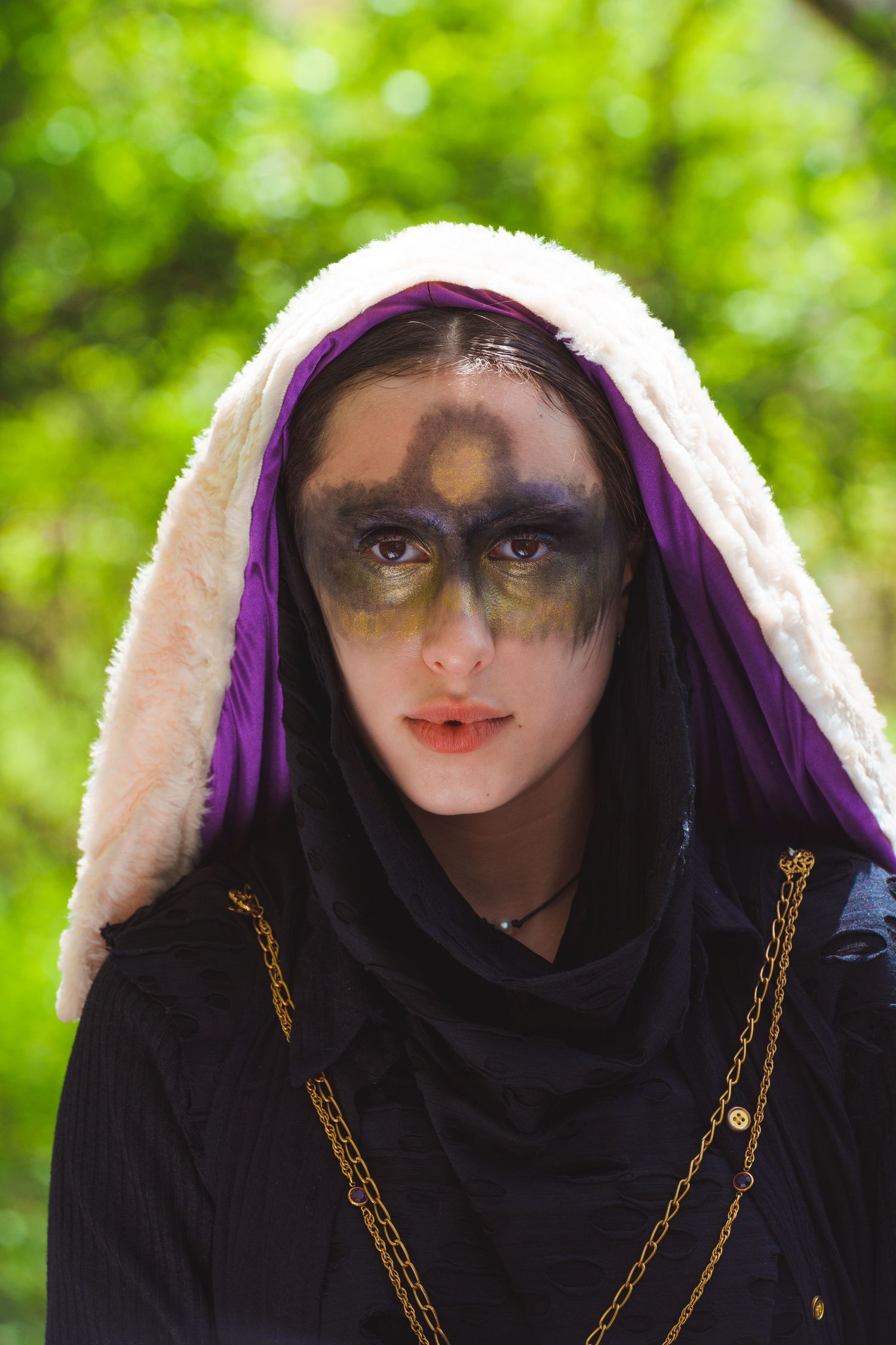 Figure wearing a dark hooded cloak with gold embellishments stands against green foliage.
