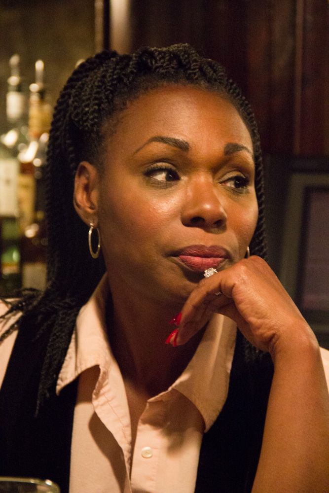 A woman with braided hair rests her chin on her hand, wearing hoop earrings and a pink shirt, in a bar setting.