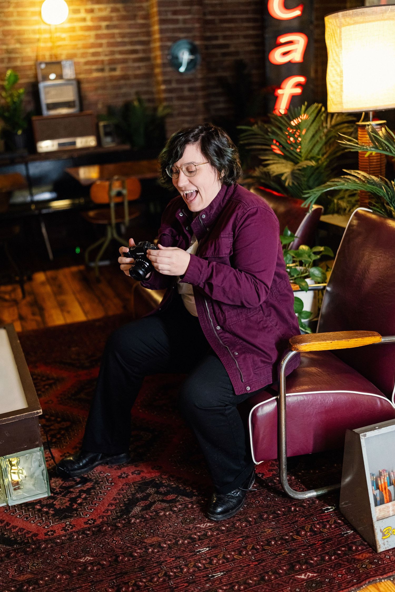 Person in purple shirt holding camera with tongue out, sitting in vintage cafe with brick walls and retro decor.