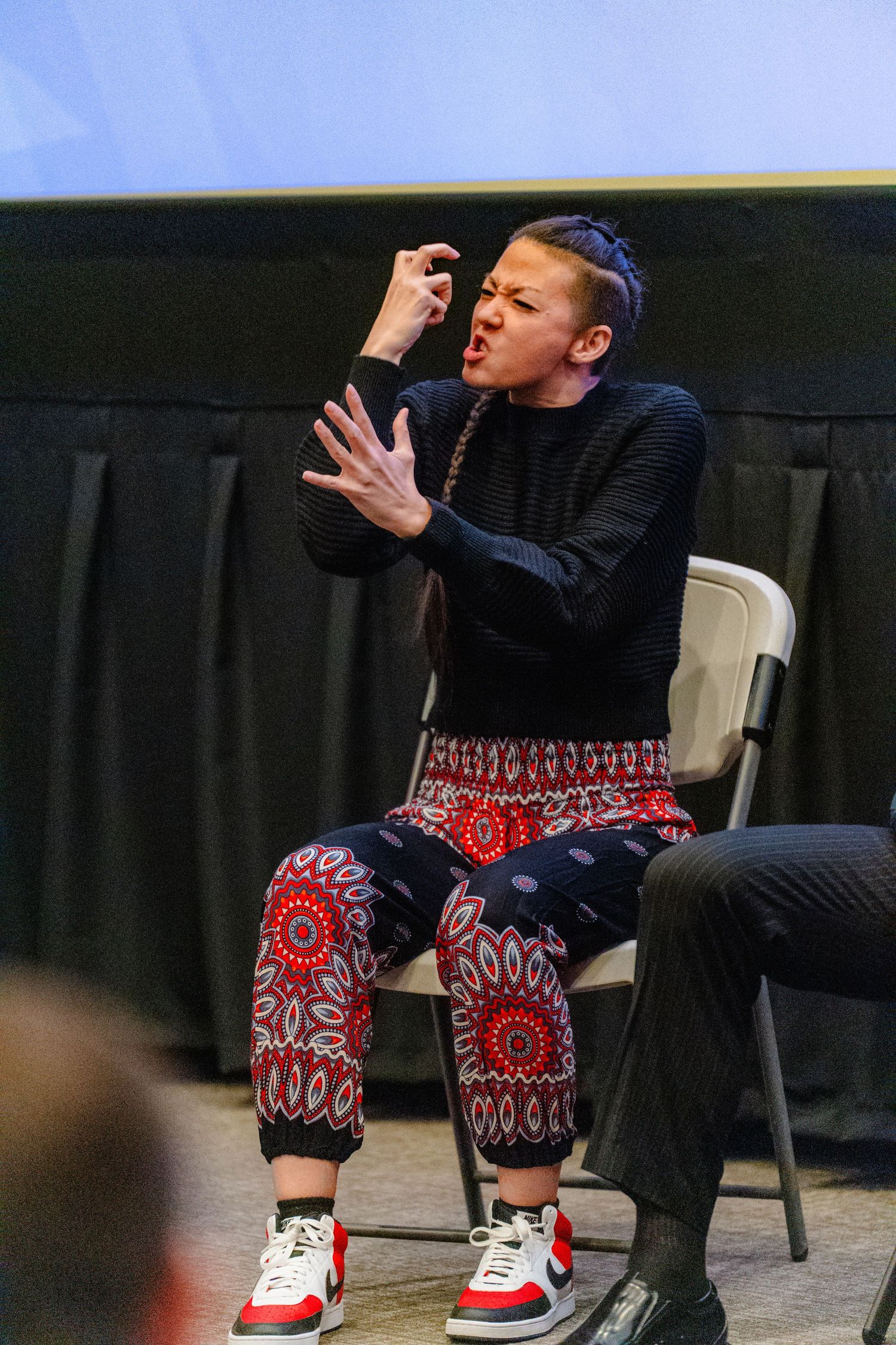 Performer in black sweater and patterned red pants gestures expressively while seated on stage during theatrical performance.