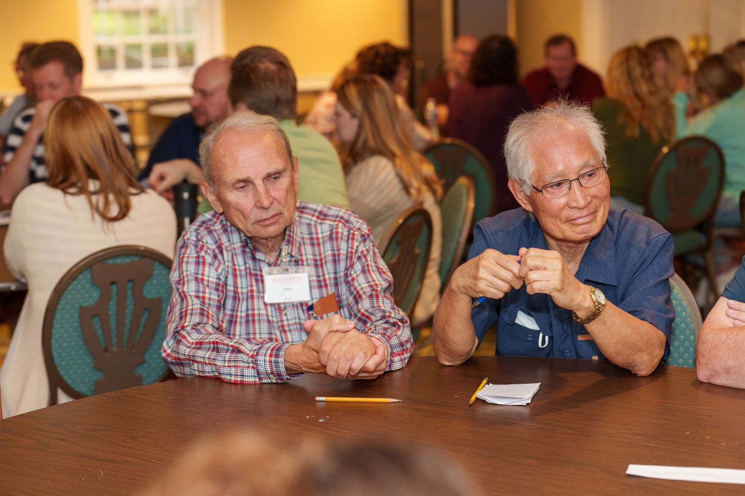 Two elderly men sit together at a table during a social gathering, engaged in conversation at an indoor event.