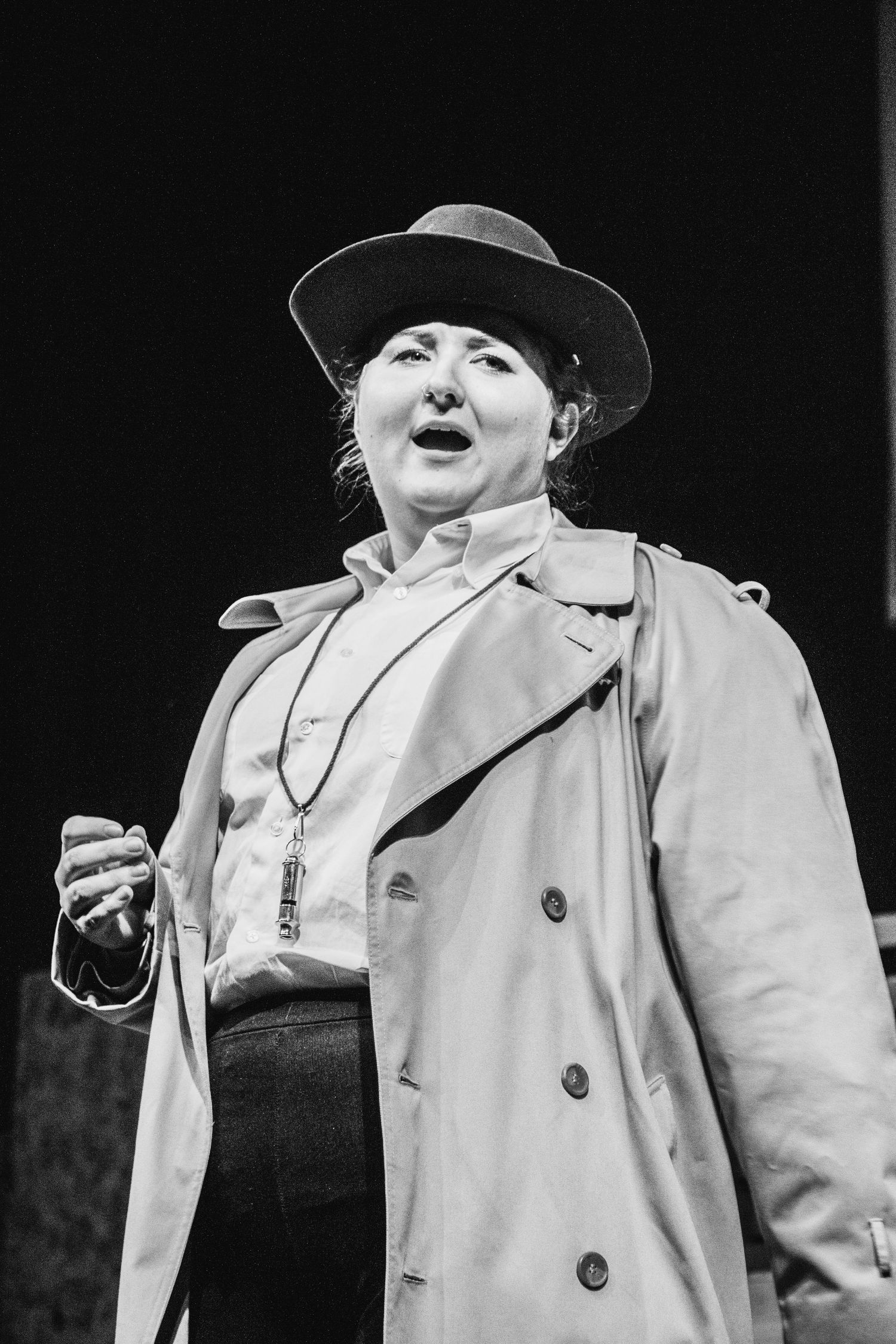 Performer in white coat and hat gesturing dramatically in black and white theatrical scene.