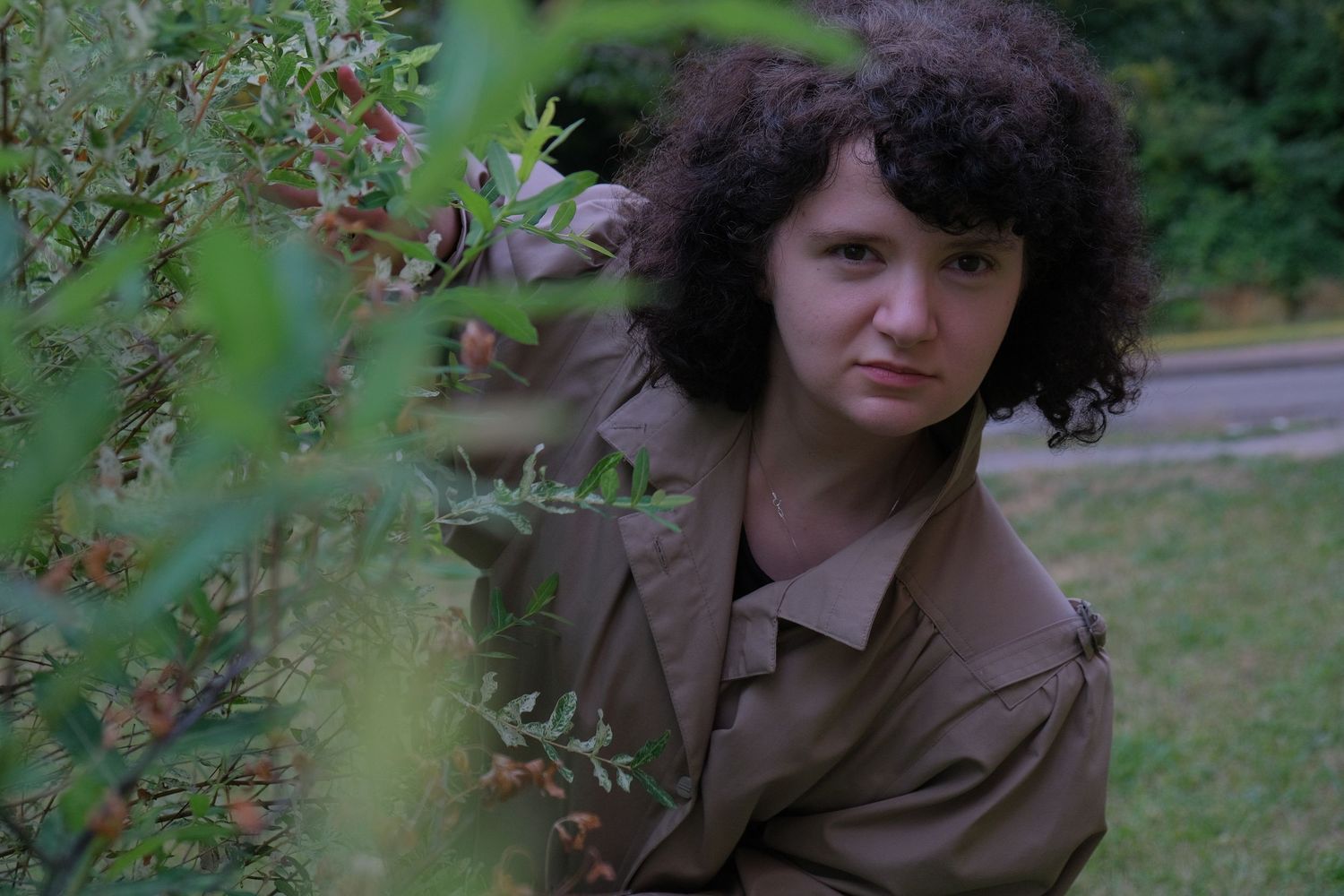Young woman with curly hair in olive jacket poses thoughtfully among green garden shrubs outdoors.