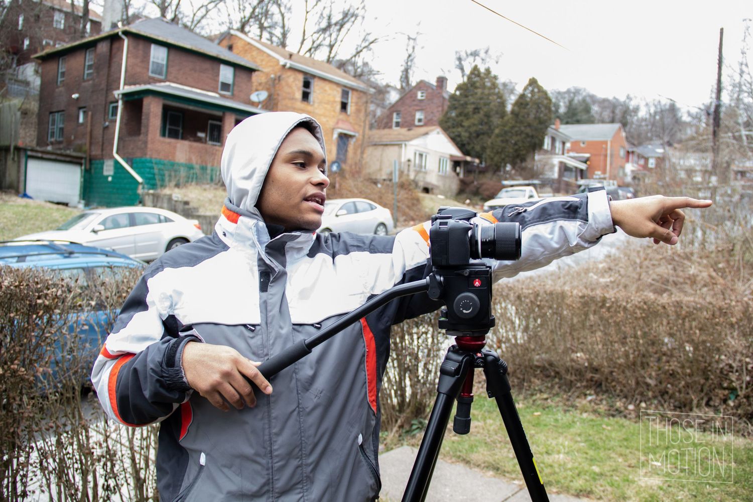 Young filmmaker directing a shot on a camera tripod outdoors in a residential neighborhood on a cloudy day.