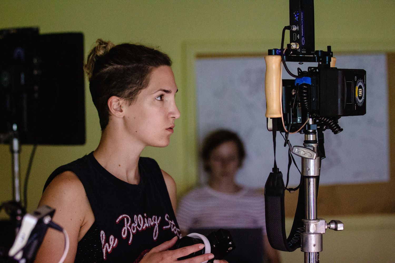 A female filmmaker in a Rolling Stones tank top monitors a professional video camera on a film set.