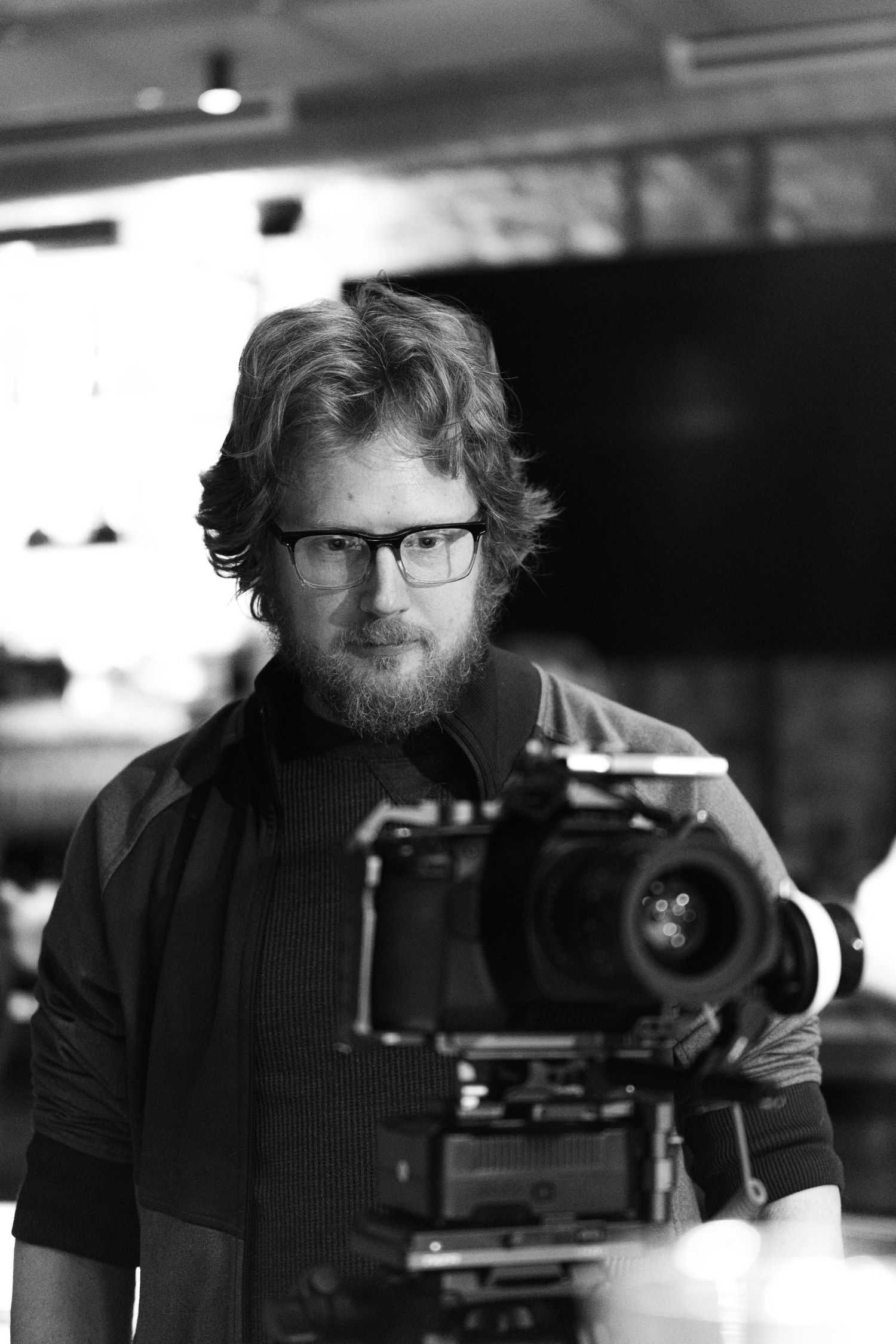 Black and white portrait of a bearded man with glasses standing behind a professional film camera on set.
