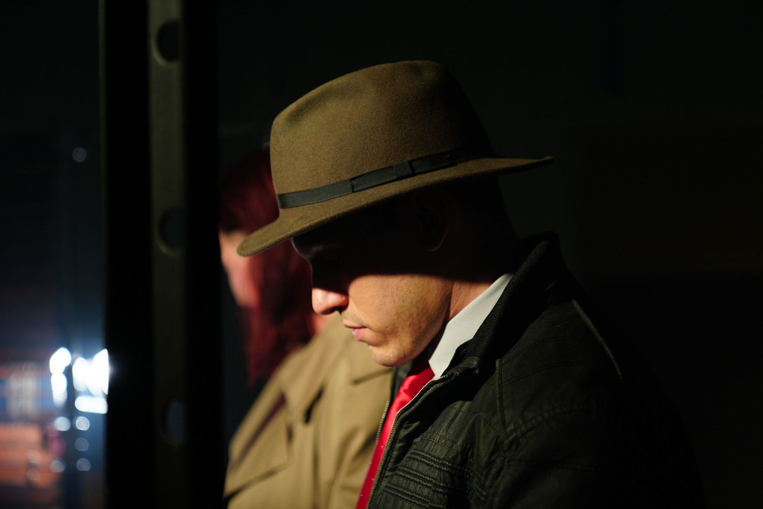 Man in vintage fedora hat and red tie standing in dramatic low light, evoking classic film noir style.