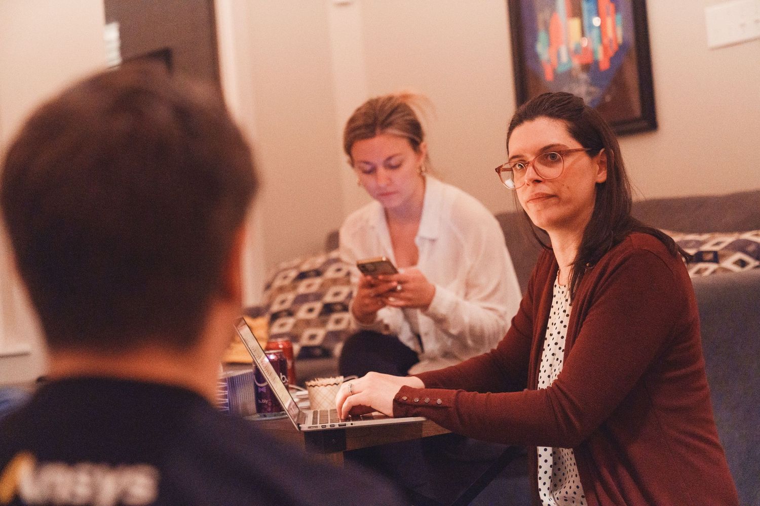 Woman with glasses works on laptop while two others collaborate in a cozy study session.