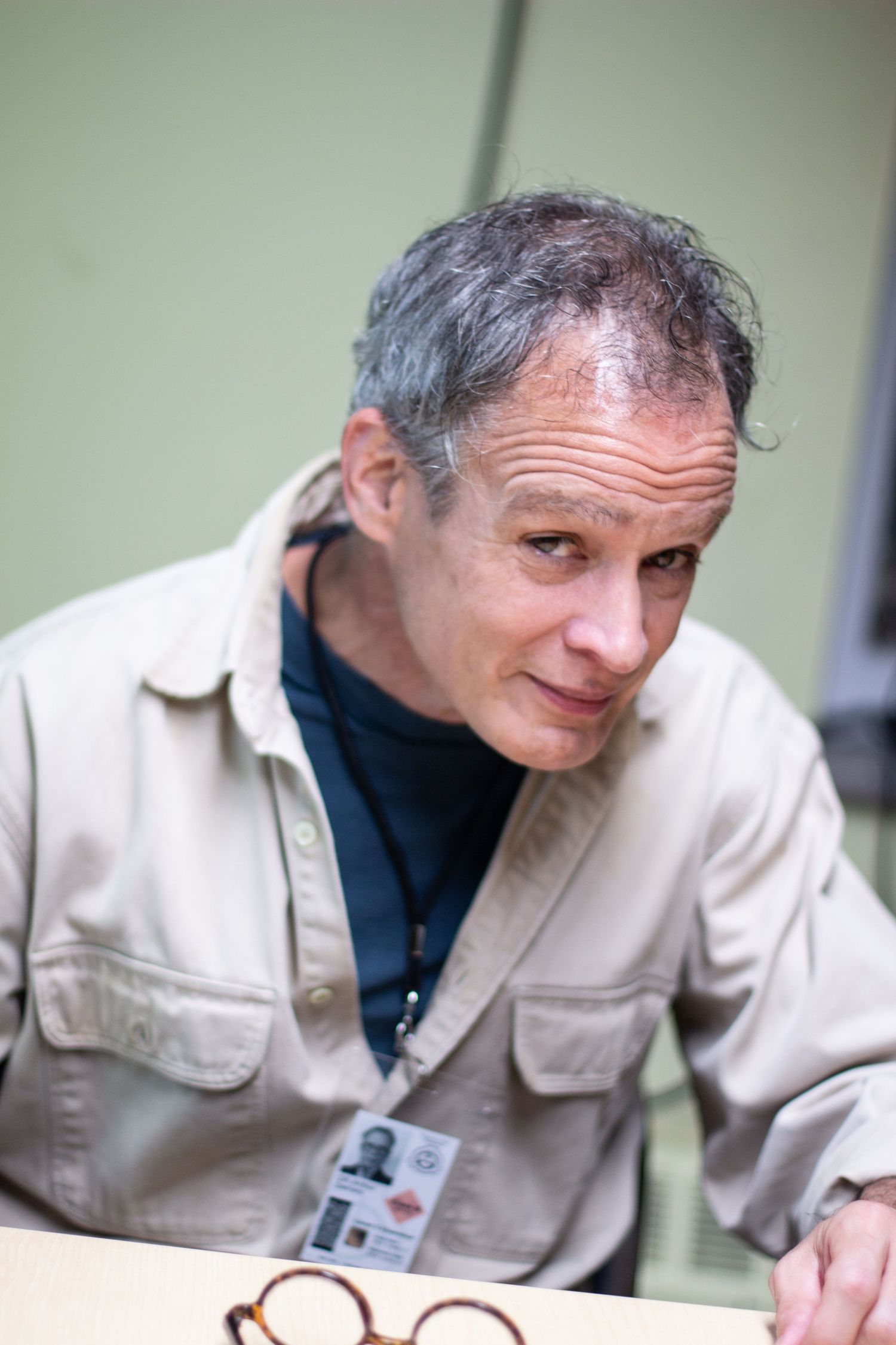 Middle-aged man with gray hair wearing a beige jacket and ID badge, sitting at a table with glasses nearby.