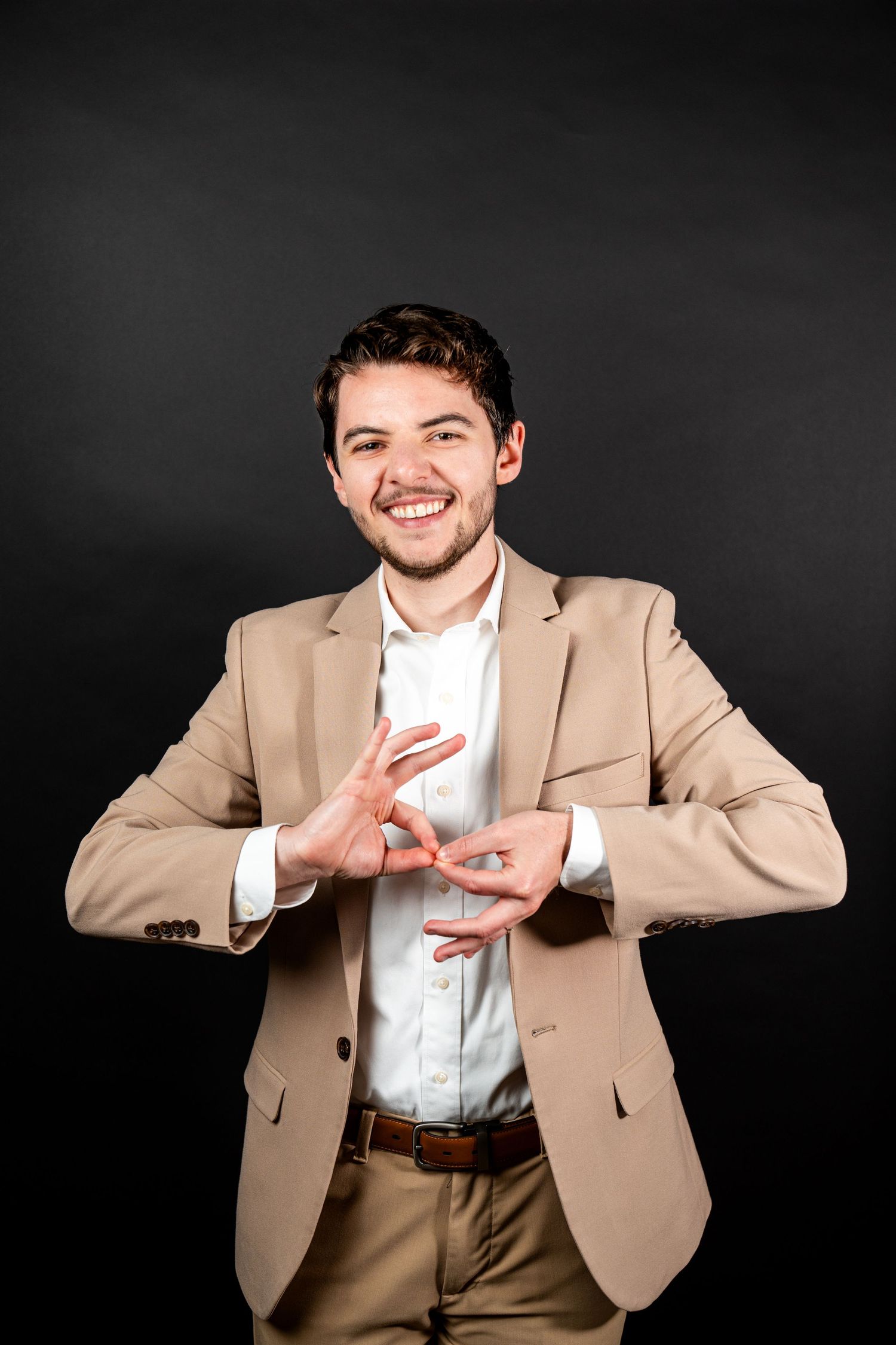 A person in a tan blazer and white shirt makes expressive gestures against a dark background.