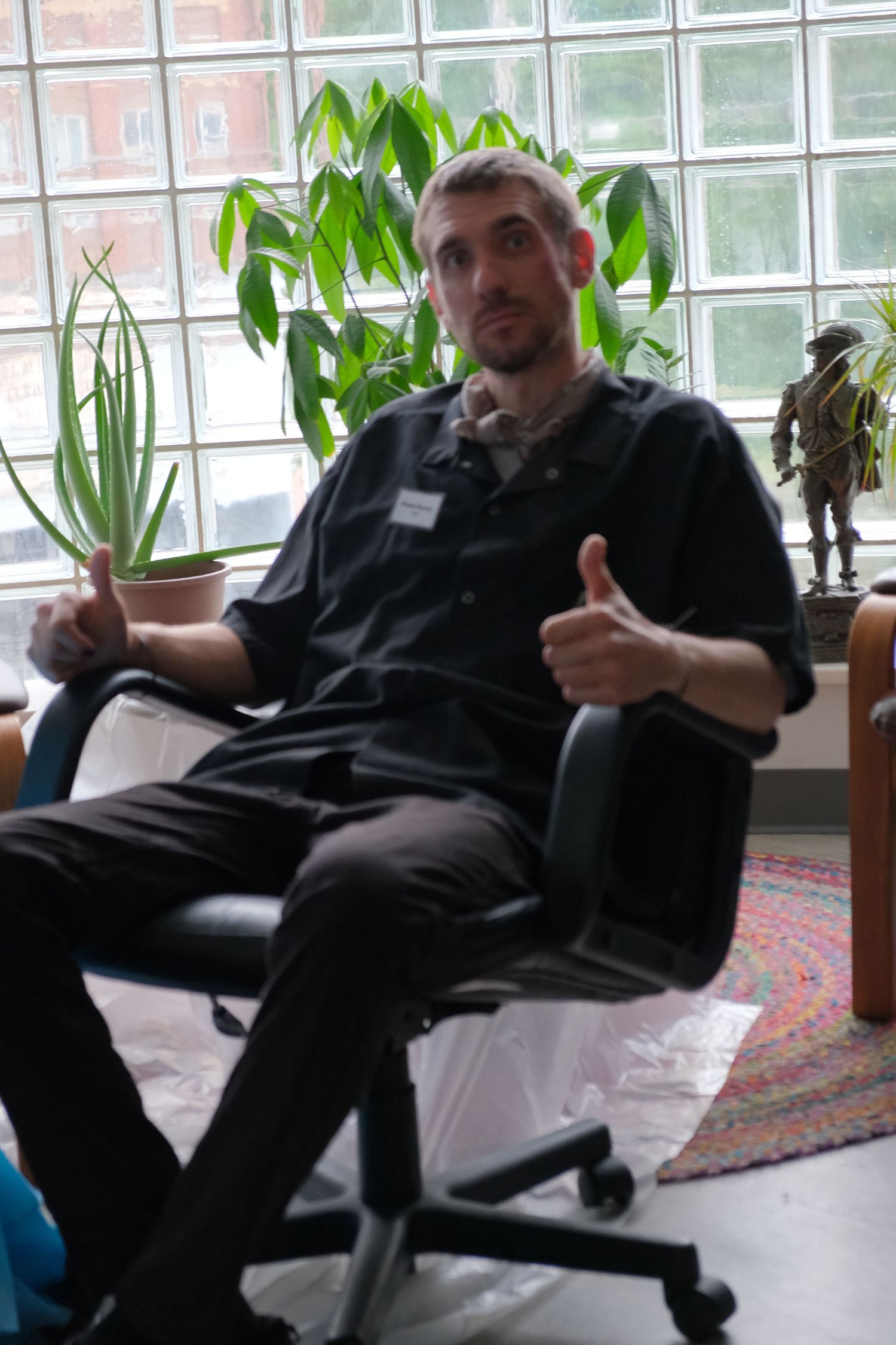 Man in black shirt sits in office chair giving thumbs up while holding a potted plant near a bright window.