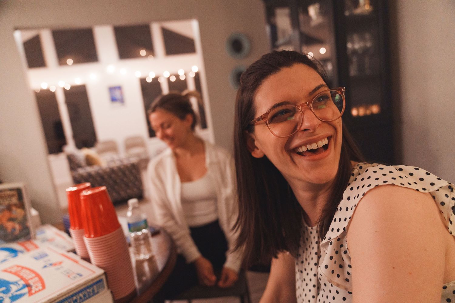 A smiling woman with glasses laughs at a casual indoor gathering with string lights in the background.