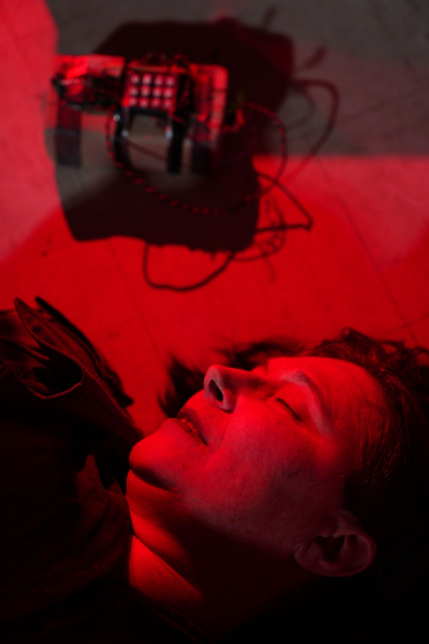 Person lying down illuminated by red light, gazing at electronic device with circuit board visible in background.