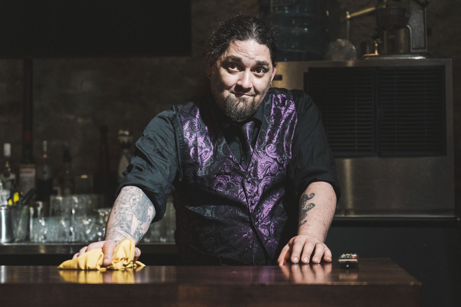 Tattooed bartender in purple vest wiping bar counter, looking at camera in dimly lit bar.
