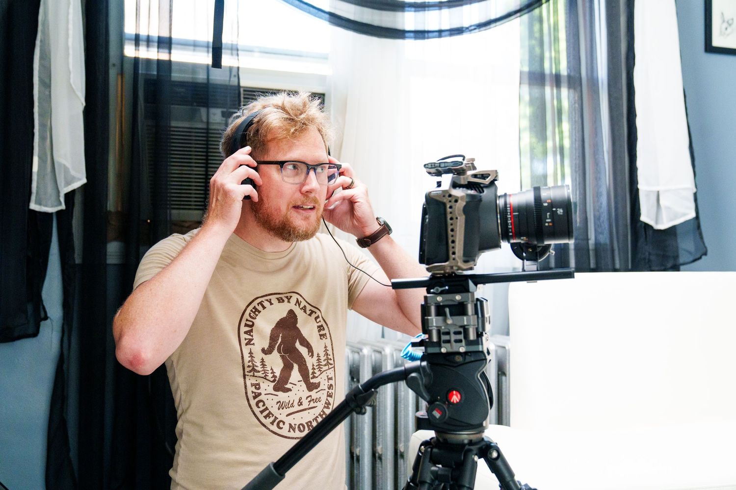 Man with glasses adjusting headphones near a professional camera on a tripod indoors.