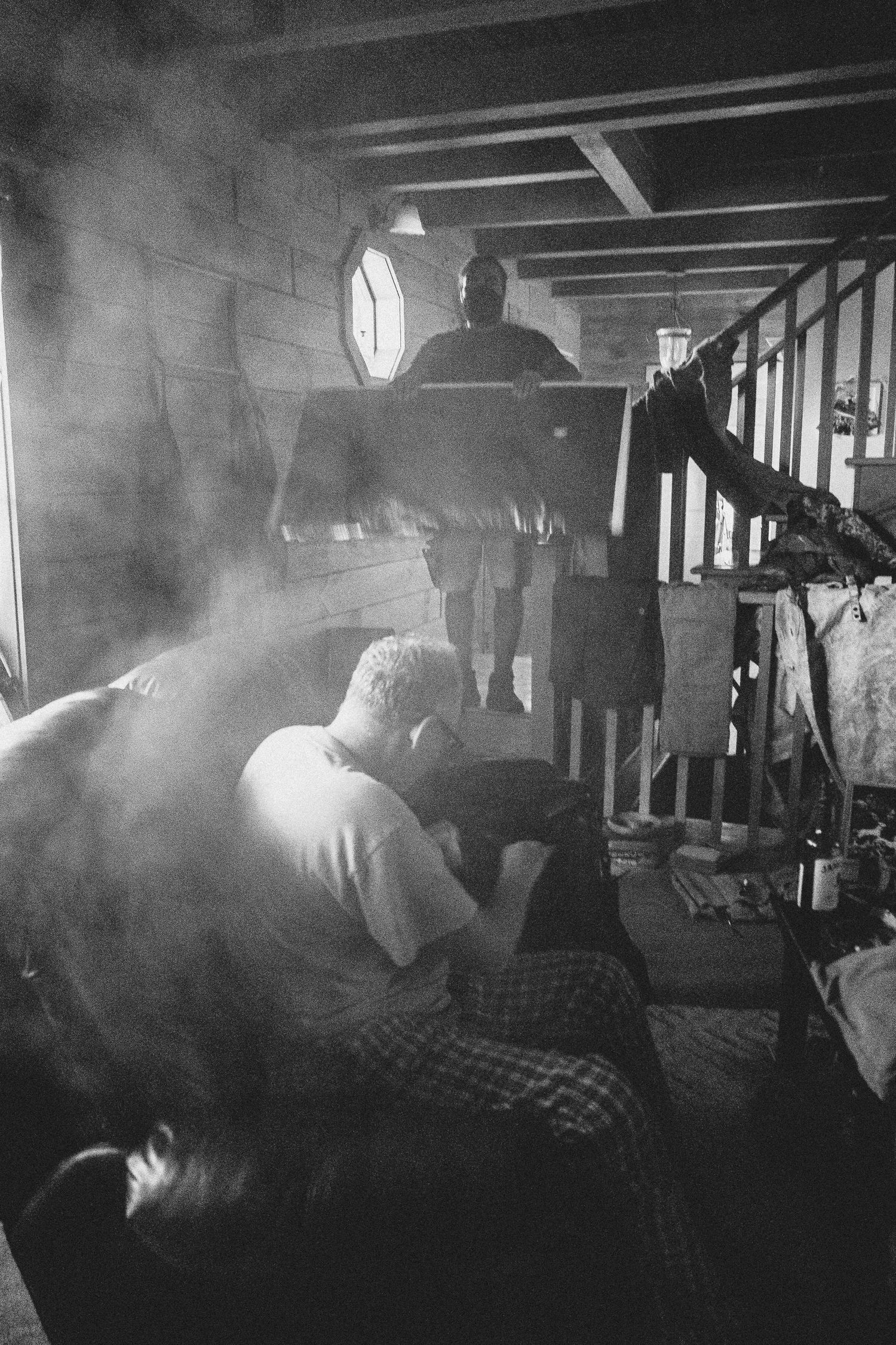 Black and white photo of men in a smoky wooden interior room with dramatic lighting.