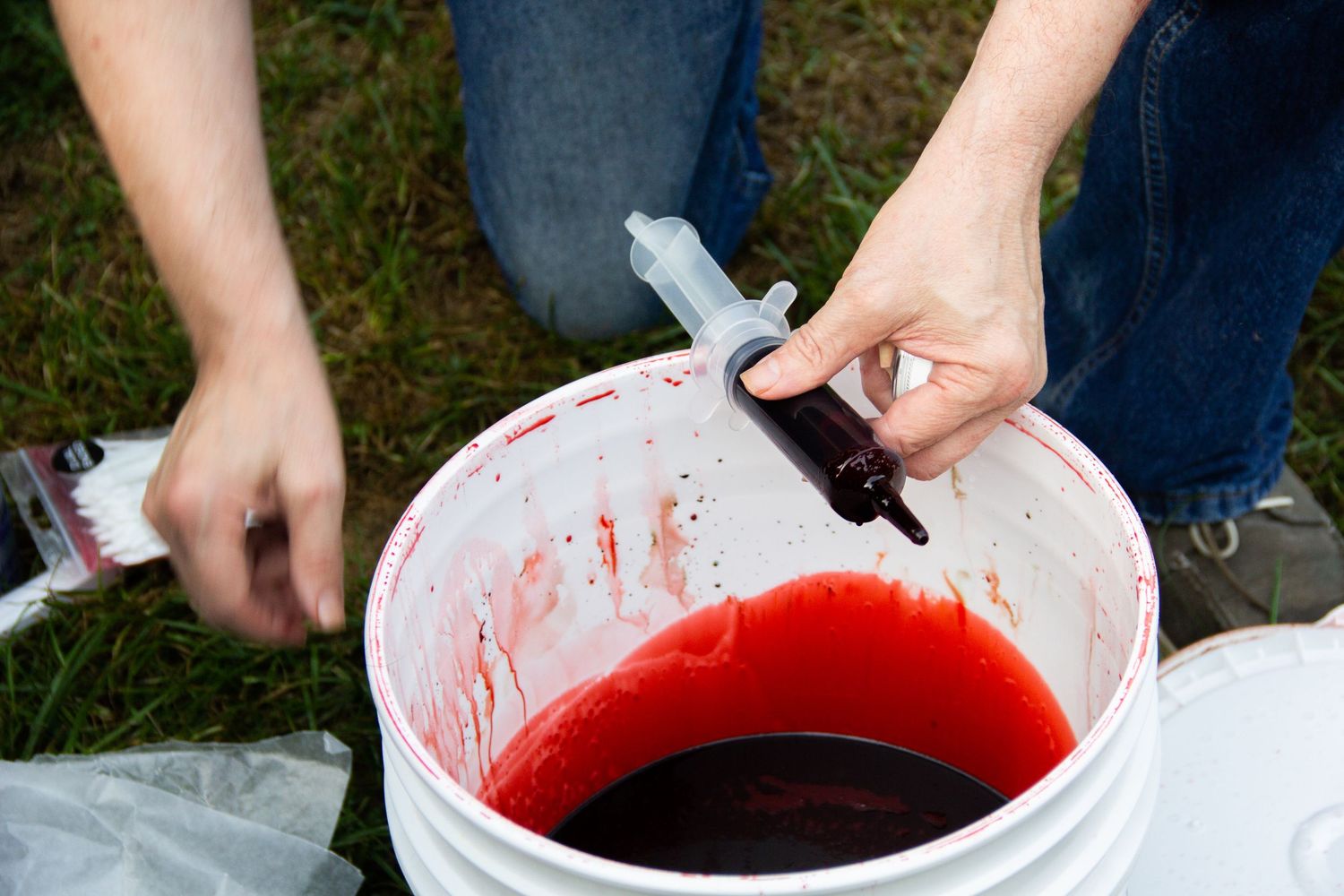 Hands filling a syringe from a bucket of red fake blood on a film production set outdoors.