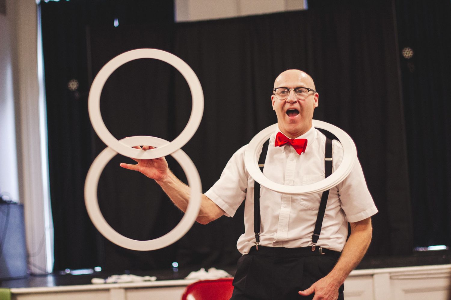 Performer in suspenders and bowtie juggles white rings on stage.