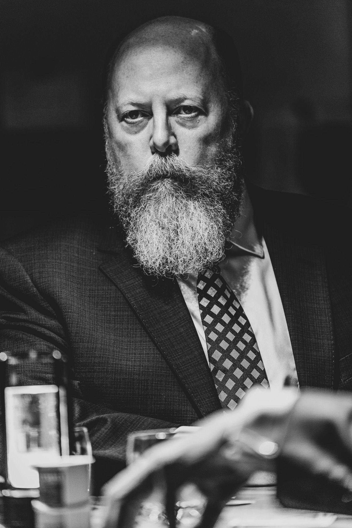 Black and white portrait of a bearded person in formal attire sitting at a dining table.
