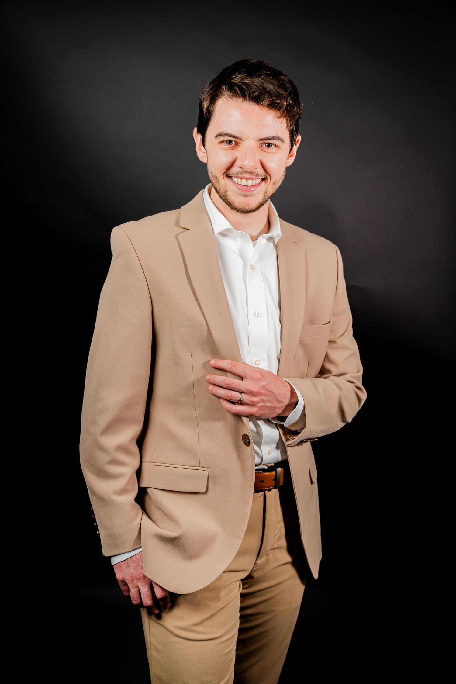 Professional portrait of someone in a tan business suit with a white dress shirt against a dark backdrop.