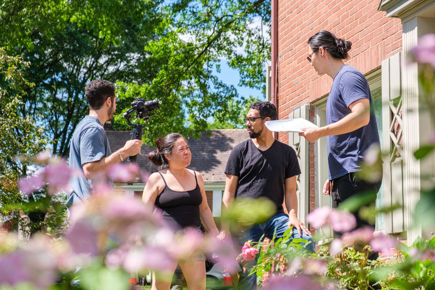 A garden scene with pink flowers and brick buildings visible in the background.