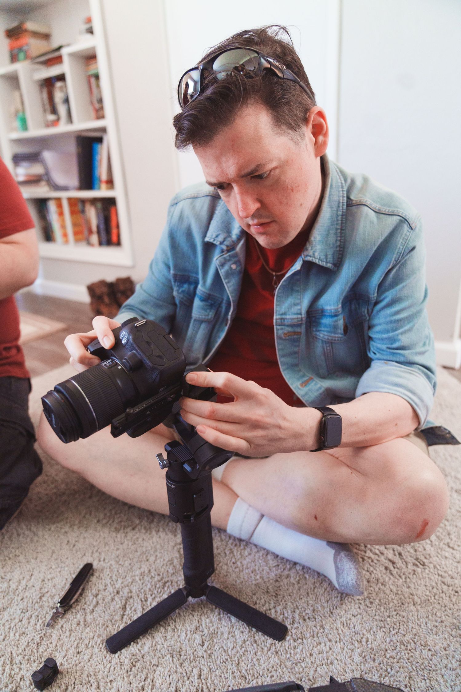 Man sitting cross-legged on carpet adjusting DSLR camera on mini tripod indoors with bookshelf in background.