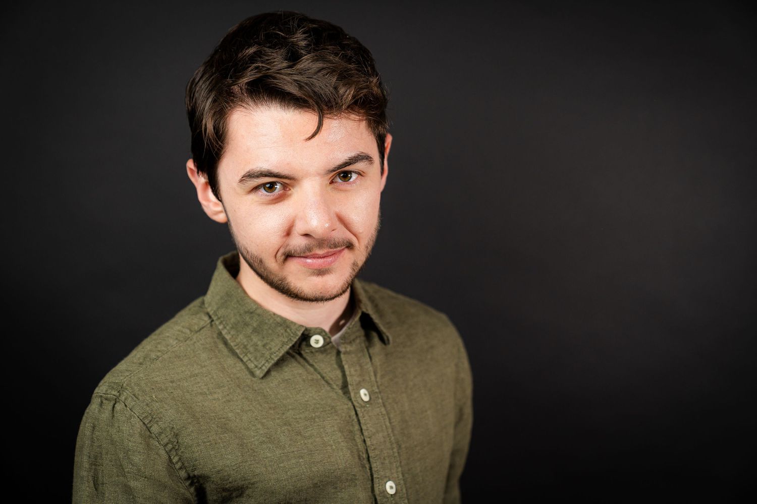 A man in an olive green button-down shirt poses against a dark background in a professional headshot series.