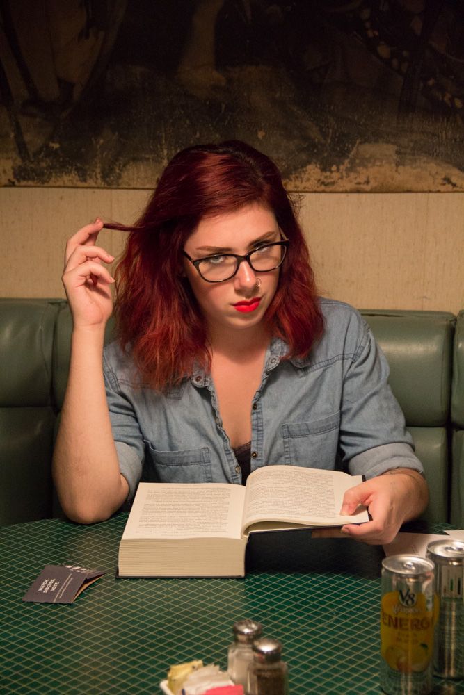 Red-haired woman with glasses reading a book at a diner booth, wearing a denim shirt with red lipstick.