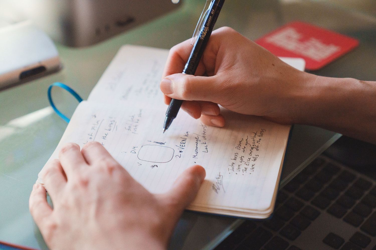 Close-up of hands writing notes and sketches in an open notebook with a black pen on a desk.