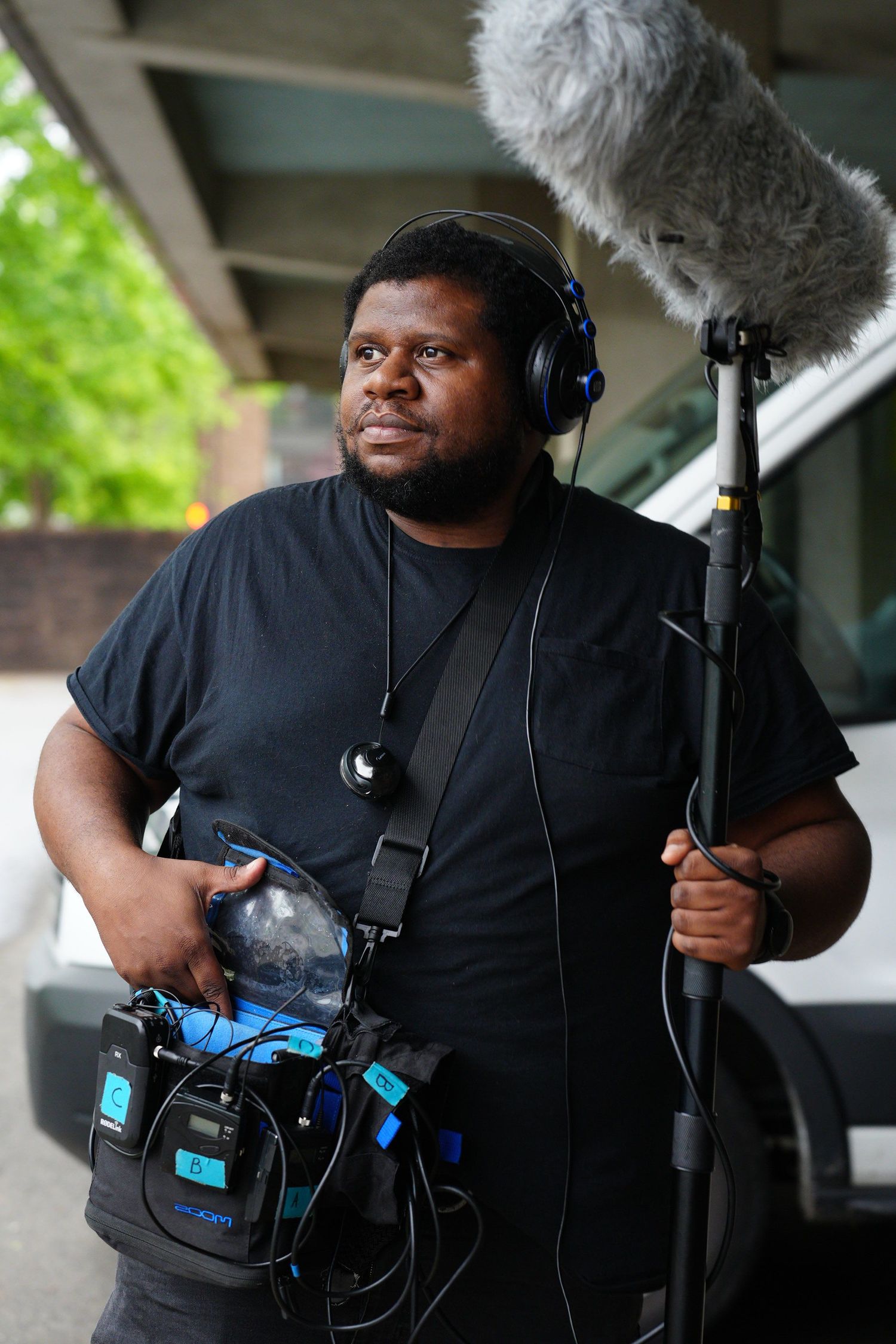 Sound technician holding boom microphone and audio equipment wearing headphones outdoors on location.