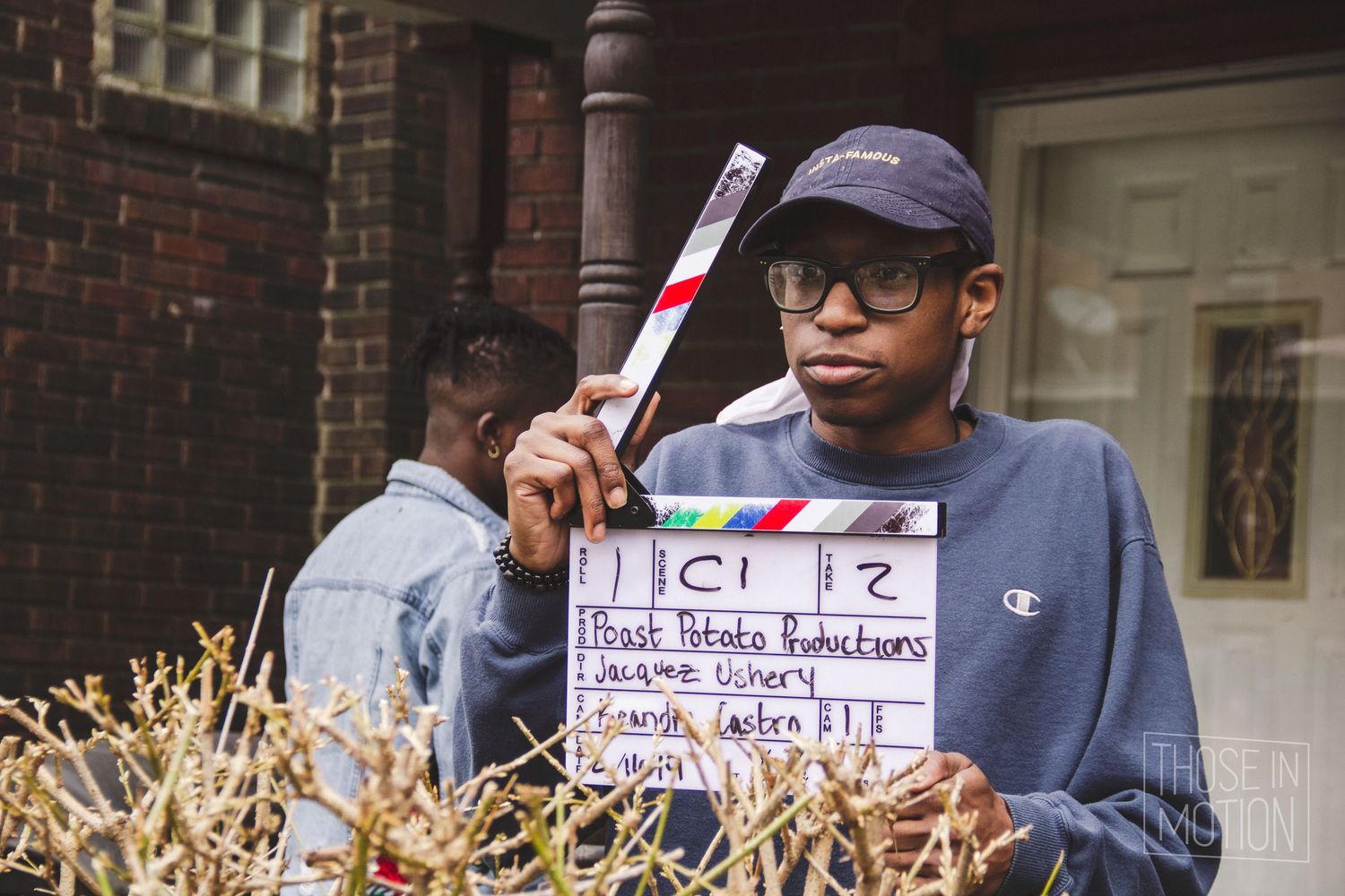 Young filmmaker holding a clapperboard on an outdoor film set, wearing a blue hoodie and cap.