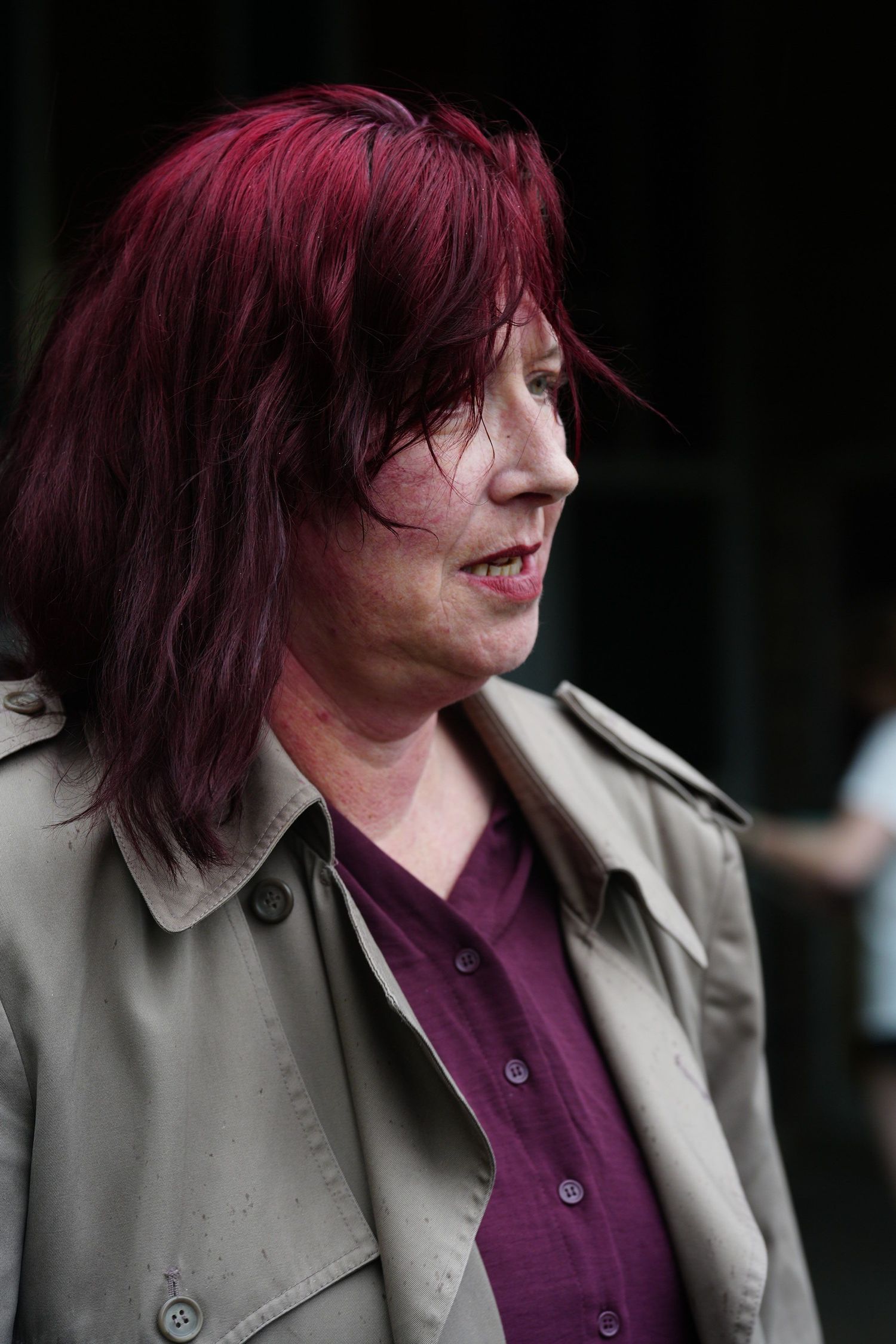 Woman with vibrant red hair wearing a beige trench coat and purple shirt, shown in profile view outdoors.