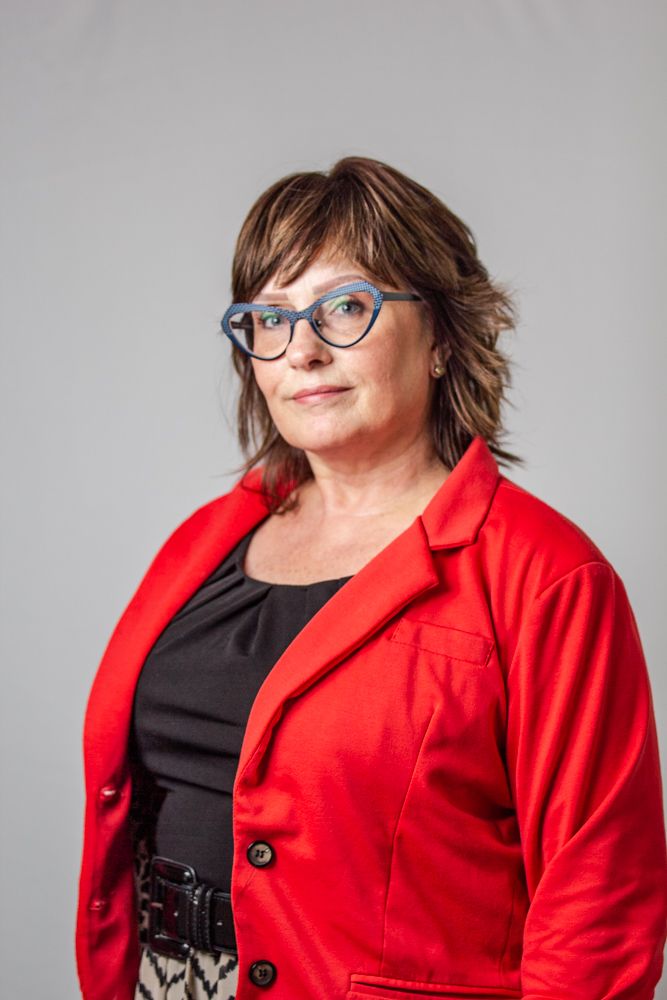 Professional portrait of someone wearing a bright red blazer, blue glasses and layered brown hair against a gray background.