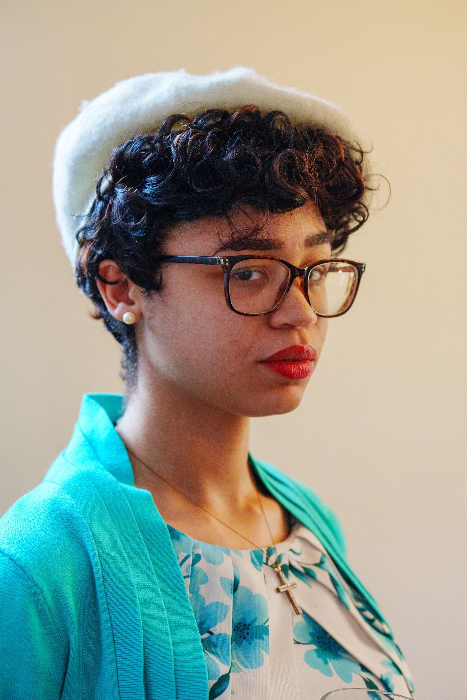 Young woman with curly hair wearing white beret, tortoise glasses, red lipstick, and teal cardigan poses for portrait.