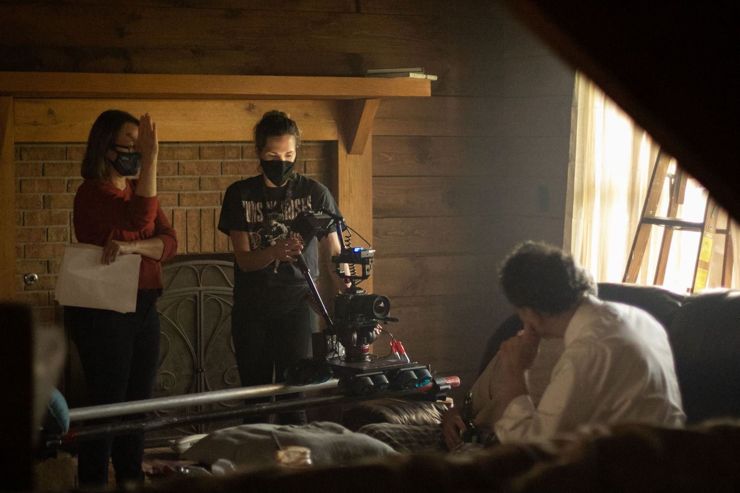 Film crew with cameras and masks work on a dimly lit cabin set with an actor seated nearby.
