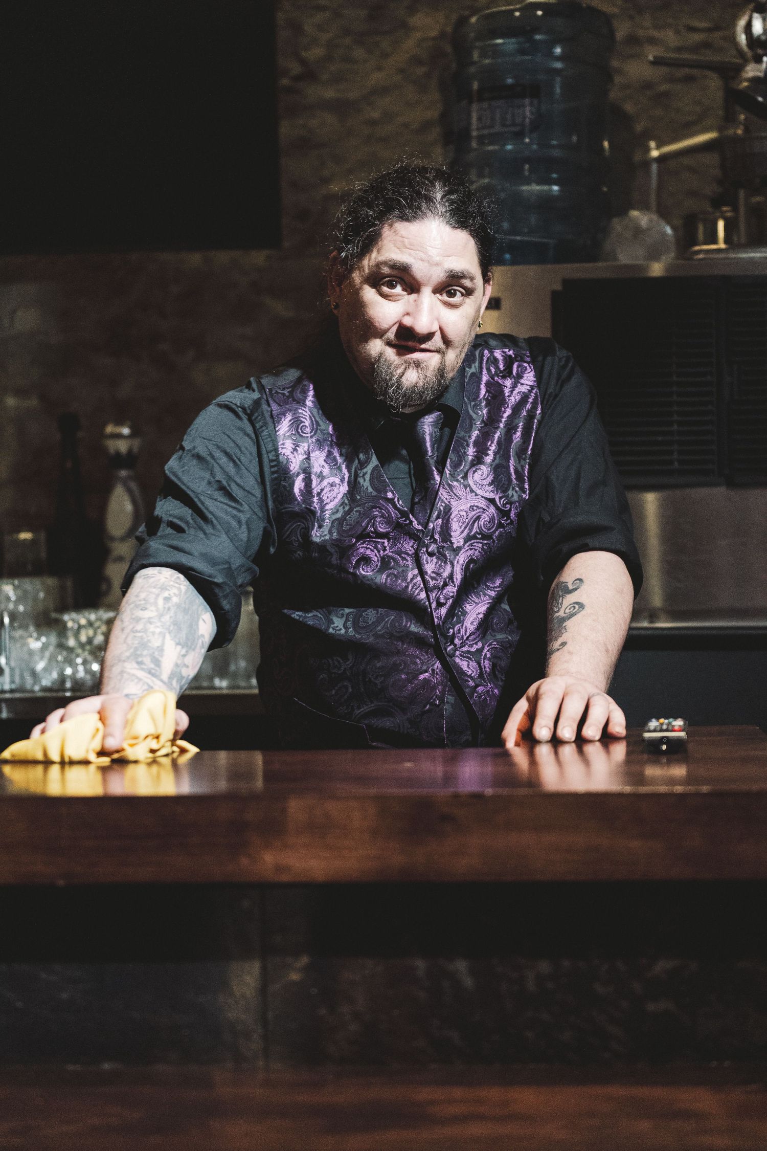 Bartender in purple vest wiping down wooden bar counter in dark atmospheric restaurant setting.