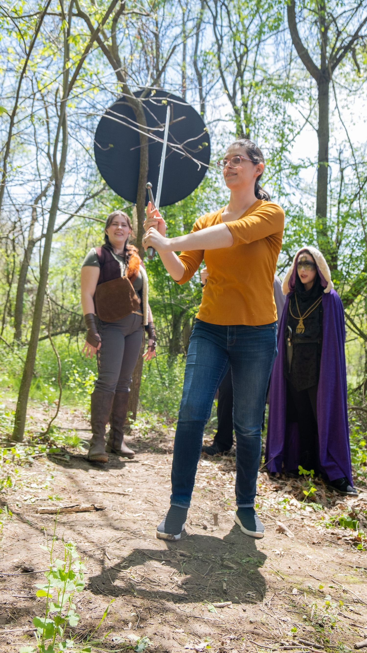 Group of performers practicing with props in a sunlit forest clearing.