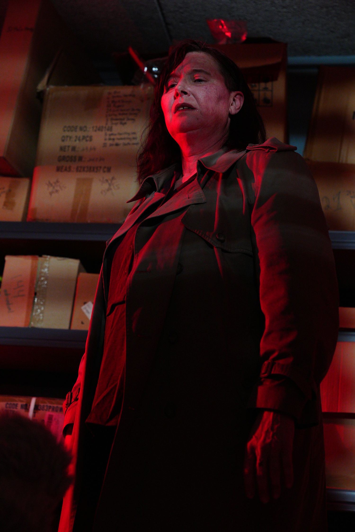 Woman in black trench coat illuminated by dramatic red lighting standing near shelves in a dark room.