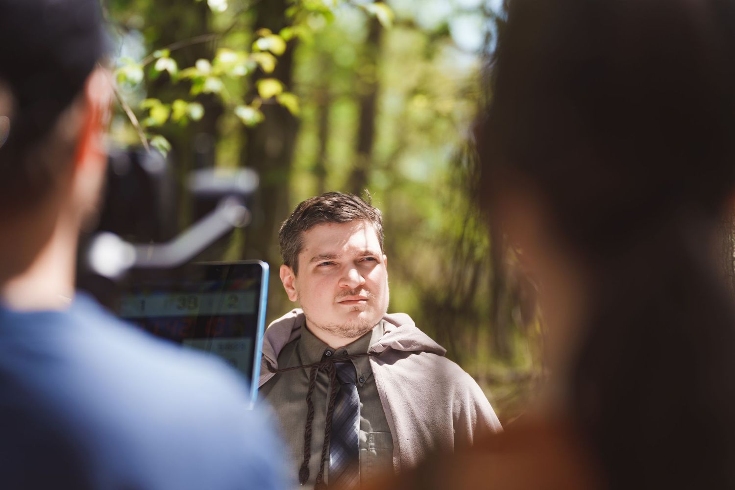 A young man sitting outdoors in sunlight, engaged in conversation with others in a wooded setting.