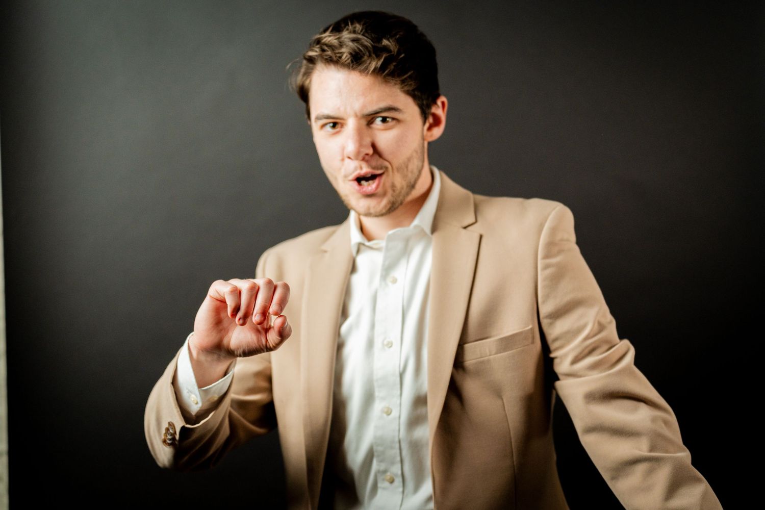 Professional studio portrait of someone in a beige suit jacket and white shirt against a dark background.