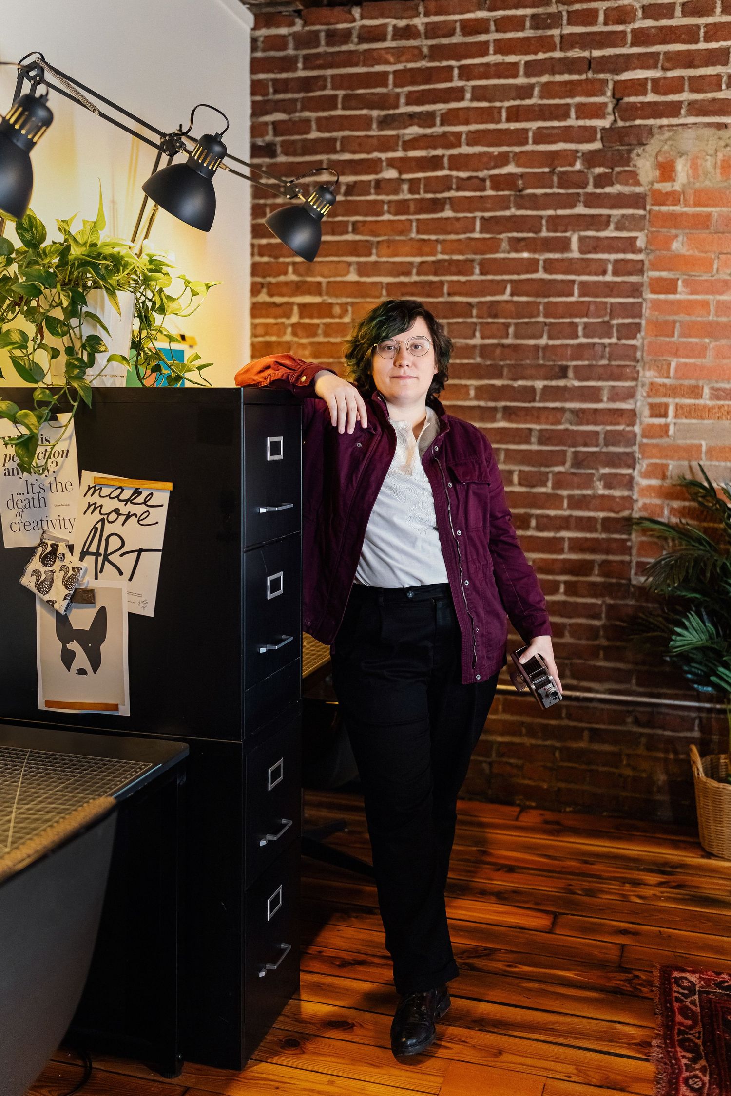 A person in a burgundy shirt leans against a black filing cabinet in a brick-walled studio with art posters and a desk lamp.