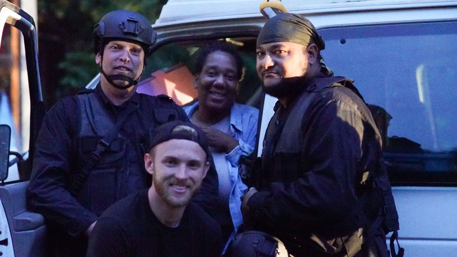 Four people pose together near a van, with two wearing tactical SWAT gear on a film set.