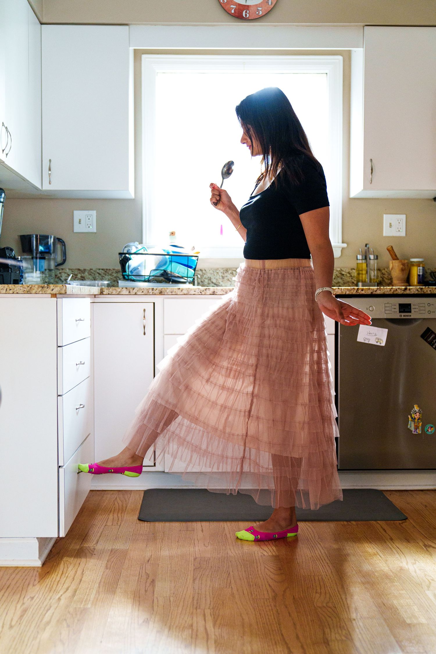 Woman in pink tulle skirt and black top tasting food while cooking in a modern white kitchen.