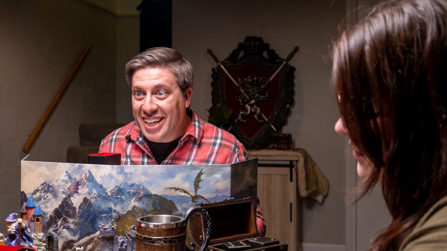 A smiling man in a red plaid shirt holds a mountain landscape painting while talking to a woman indoors.