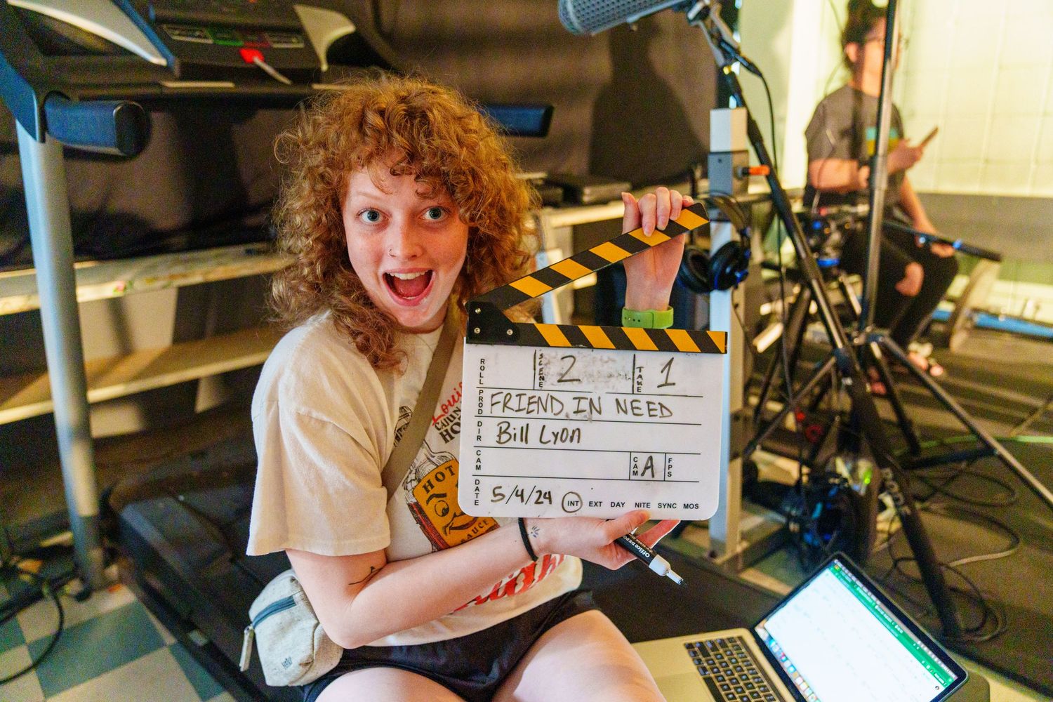 Curly-haired person smiles holding a film clapperboard labeled Friend In Need on a movie set.